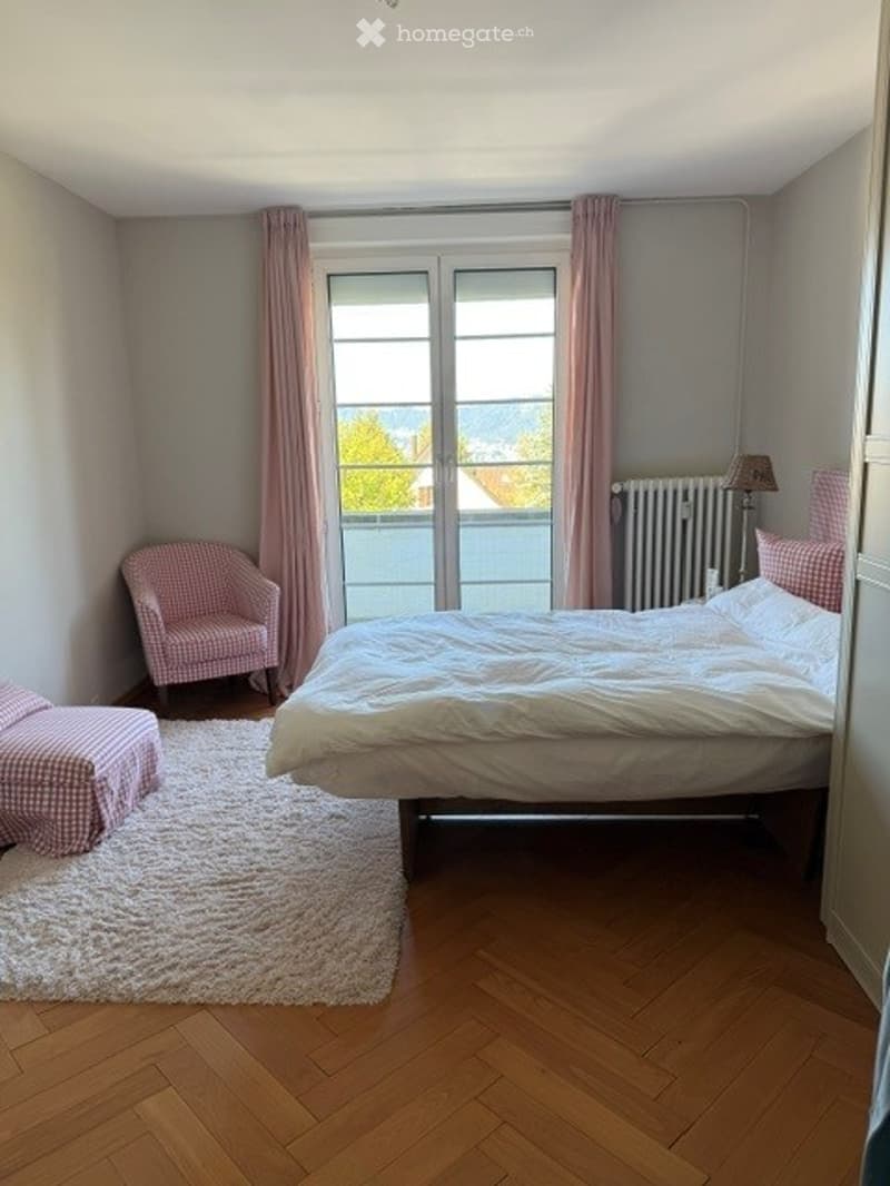 Cozy bedroom with wooden flooring, a large bed with white bedding, a pink armchair, a matching pink stool, and a radiator near the window with pink curtains.