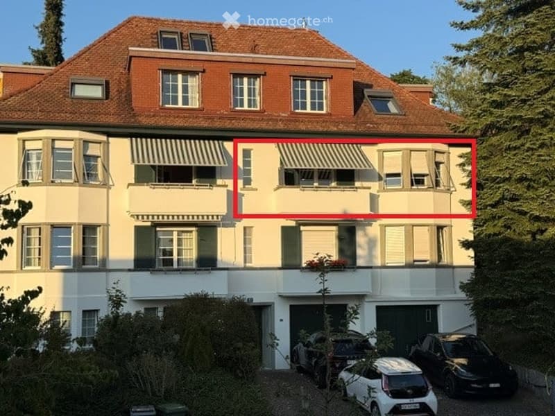 Multi-story residential building with a red-tiled roof, multiple windows, balconies, and a garage with parked cars.