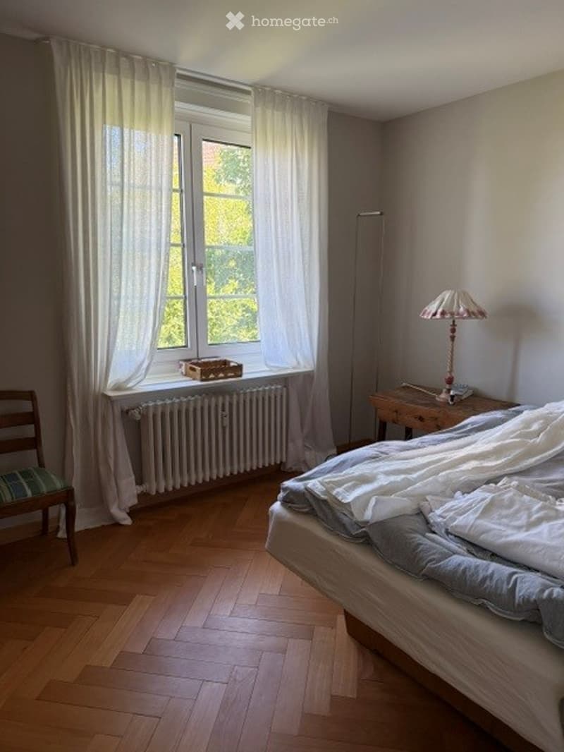 Cozy bedroom with wooden flooring, large window with white curtains, bed with white and grey bedding, wooden chair, small table with lamp, and radiator below the window.