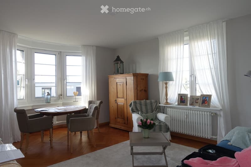 Bright living room with wooden flooring, large windows with white curtains, a sofa, armchair, coffee table, dining table with chairs, a wooden cabinet, and decorative items including framed photos and a flower vase.