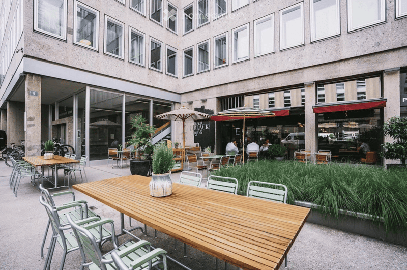 Courtyard seating area with wooden tables, green chairs, umbrellas, and potted plants, adjacent to a modern building with large windows.