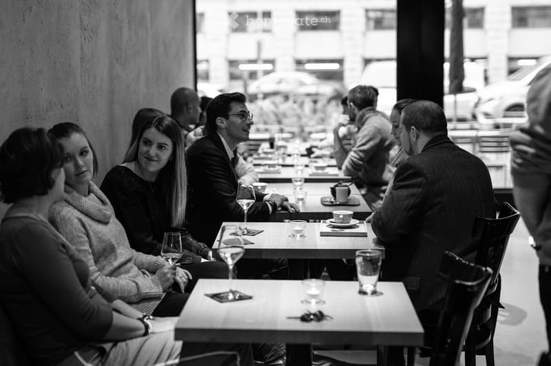 A group of people seated at tables in a restaurant or cafe, enjoying drinks and conversation. Tables are set with wine glasses, cups, and napkins.