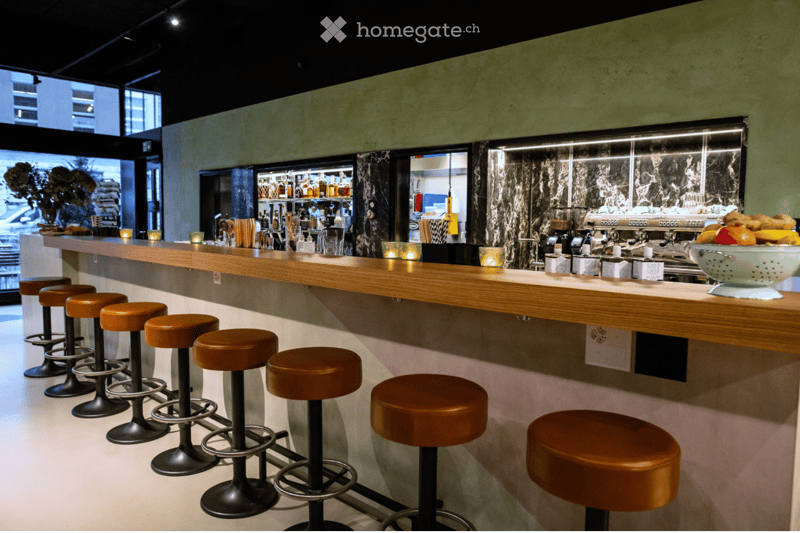 Modern bar area with a wooden counter, multiple bar stools, a refrigerator, a coffee machine, and a bowl of fruit on the counter.