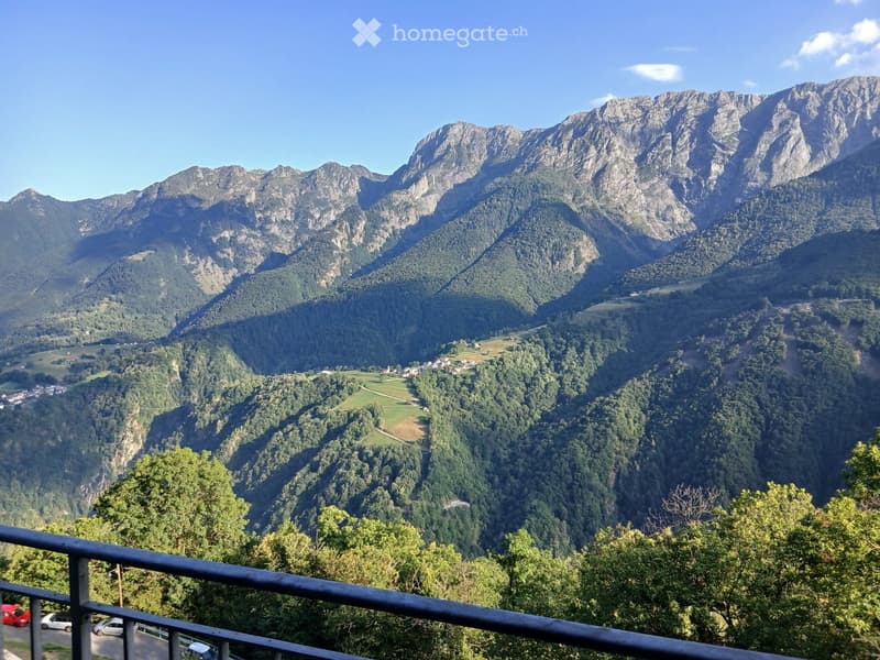 Mountain range, lush greenery, blue sky, black fence, parked cars