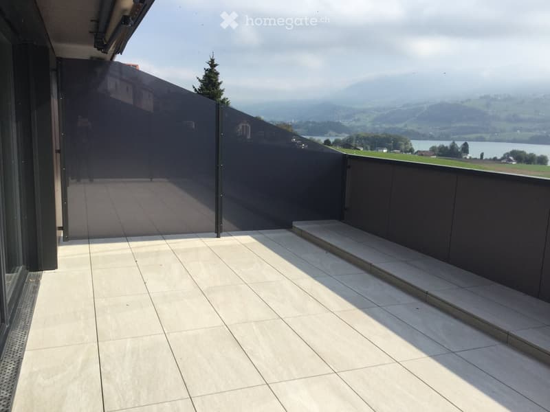 Modern balcony with light-colored tiled flooring, dark privacy screen, and scenic mountain and lake view.