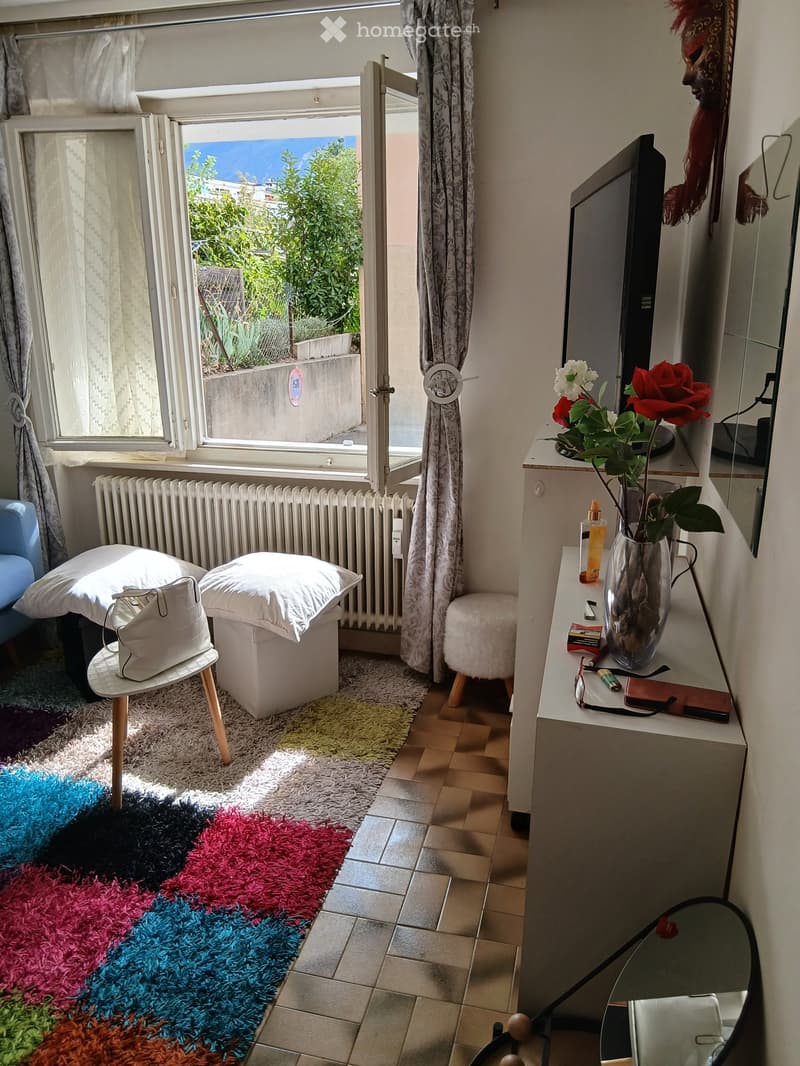 A cozy, well-lit room with a colorful shag rug, a small sofa, a white ottoman, a side table with a handbag, a vanity with a vase of flowers, and a television on a stand.