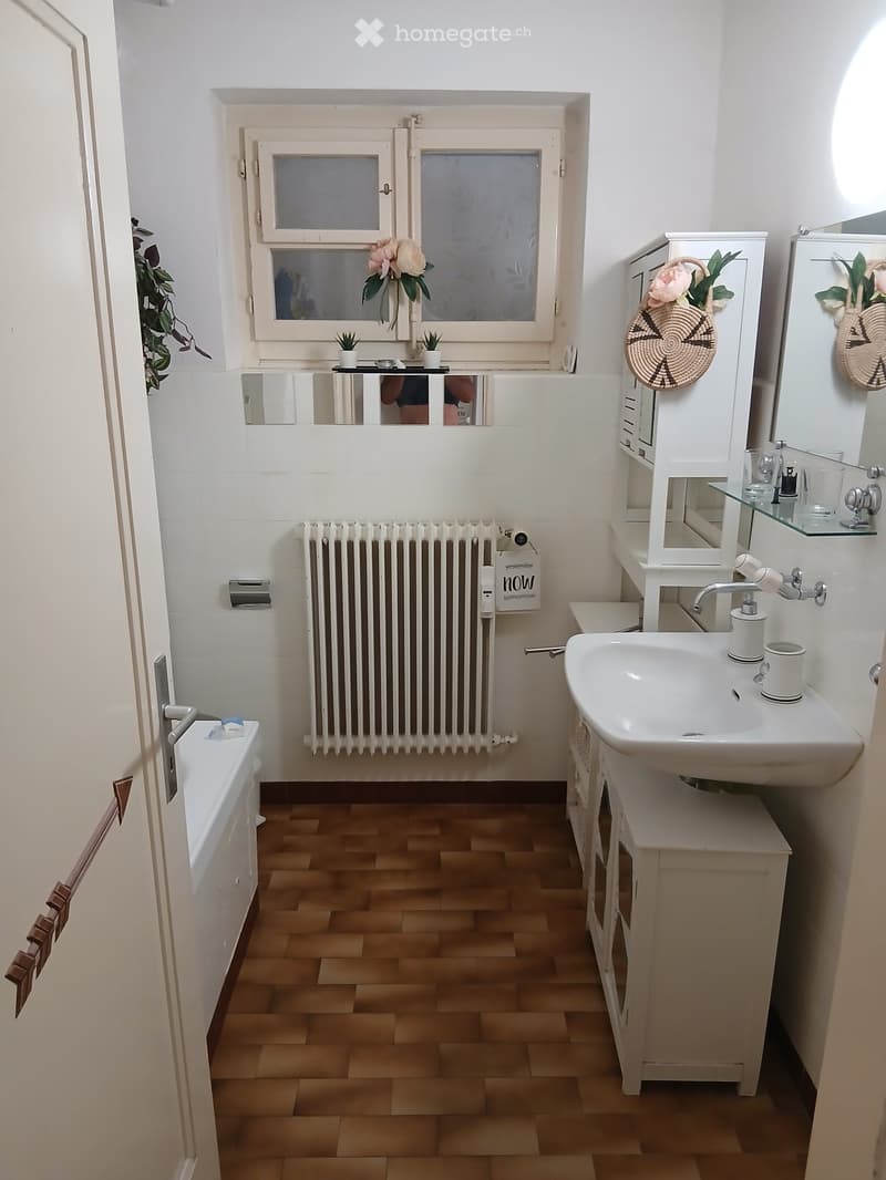 Modern bathroom with white tiles, wooden flooring, sink, mirror, radiator, and bathtub.