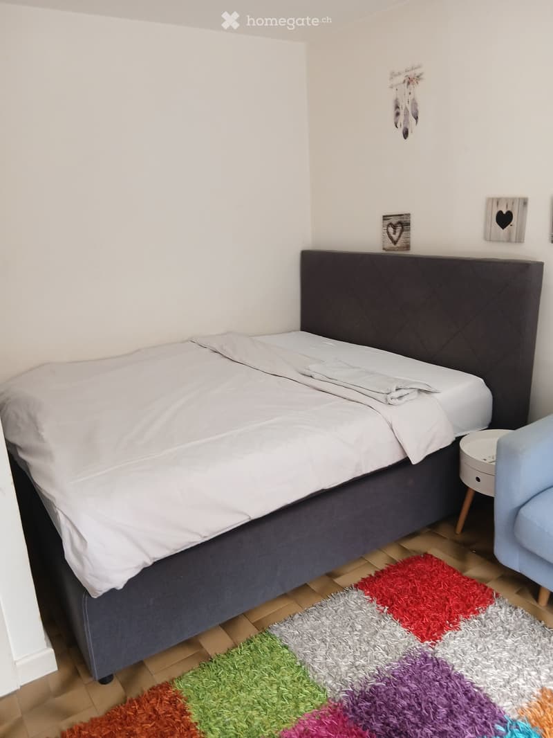 A neatly made double bed with white bedding and a dark headboard, positioned against a white wall. A colorful shag rug lies on the wooden floor, and a small round nightstand with a lamp sits beside the bed, accompanied by a light blue armchair.
