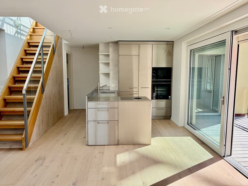Modern kitchen with light wood flooring, a staircase leading upwards, a central island with sink and faucet, built-in cabinets, and an oven. Large glass doors open to a balcony or outdoor area.