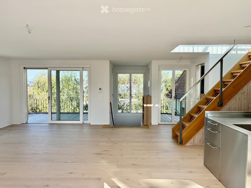 Bright, open-plan living space with light wooden flooring, white walls, large glass doors leading to a balcony, and a modern staircase leading to an upper level.