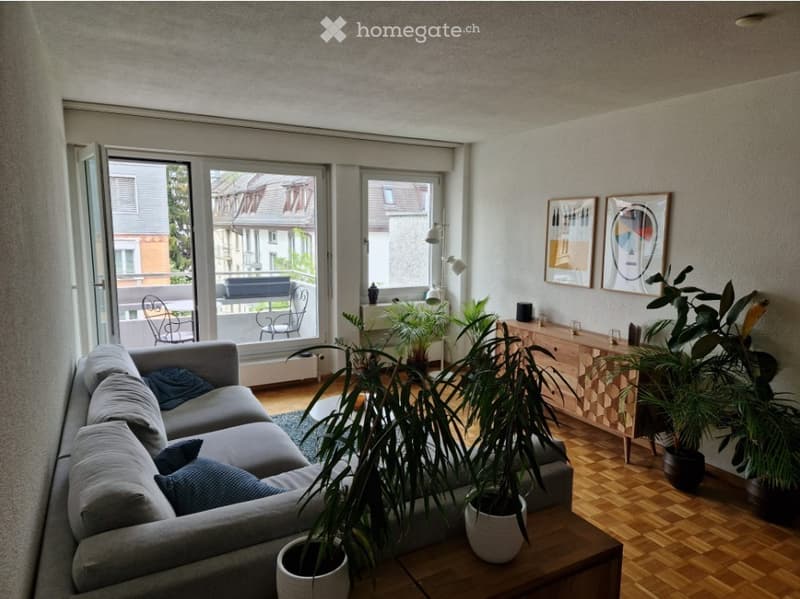Modern living room with a grey sectional sofa, coffee table, side table, potted plants, and a balcony with outdoor seating.