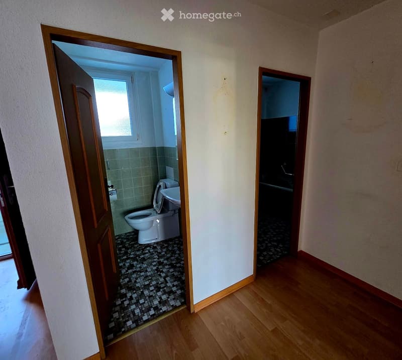 Empty hallway with wooden flooring, leading to a bathroom with a toilet, sink, and window, and another room with dark flooring.