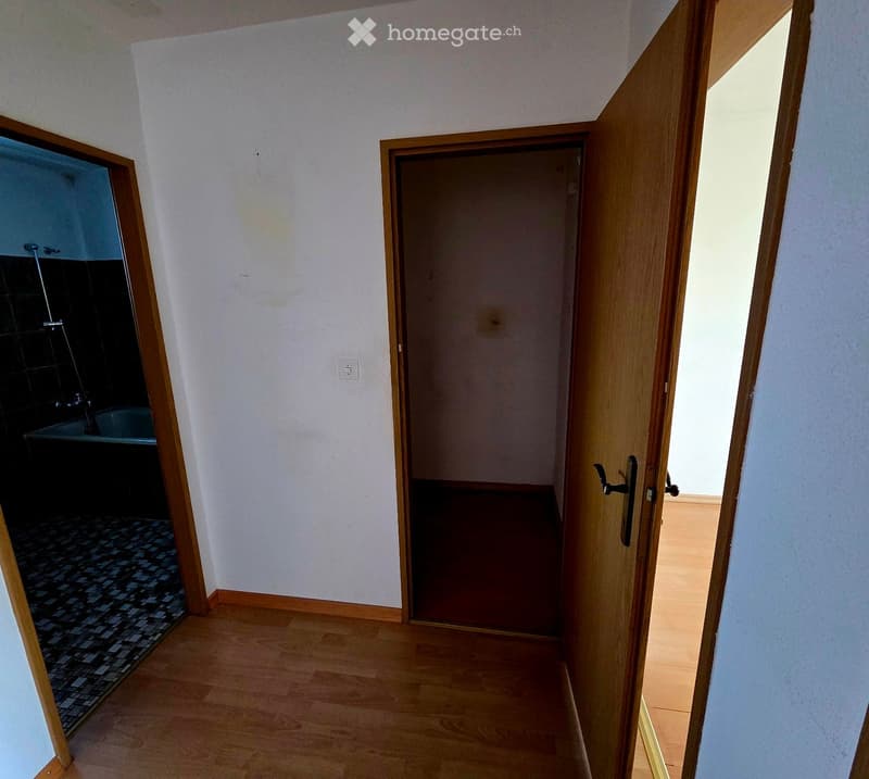 A narrow hallway with wooden flooring and white walls, featuring an open door leading to a room and a partially visible bathroom with a bathtub and tiled walls.