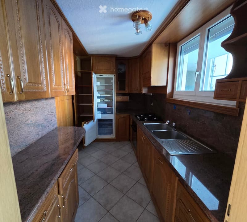 A well-lit kitchen with wooden cabinets, a stainless steel sink, a refrigerator, and a stove. The countertops are made of dark stone, and the floor is tiled.