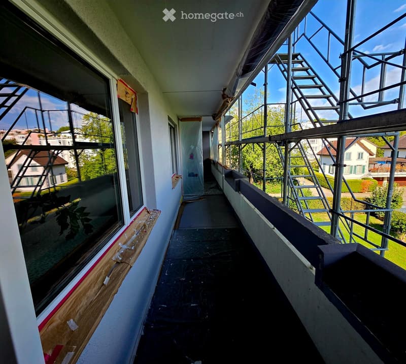 Balcony with metal railings, windows, and a view of the surrounding area. The floor is covered with protective material, and there are some construction materials visible.