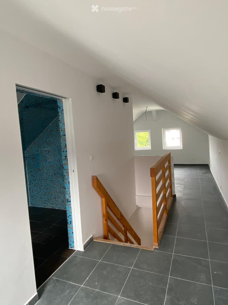 A modern hallway with dark tiled flooring, white walls, and wooden railings leading to a room with windows and a blue-tiled wall visible in the background.