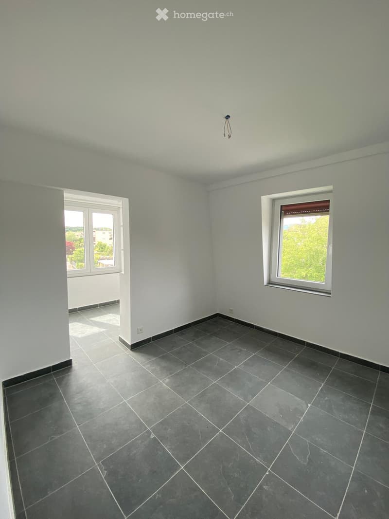 Empty room with light grey walls, dark grey tiled floor, and two windows allowing natural light. No furniture or equipment present.