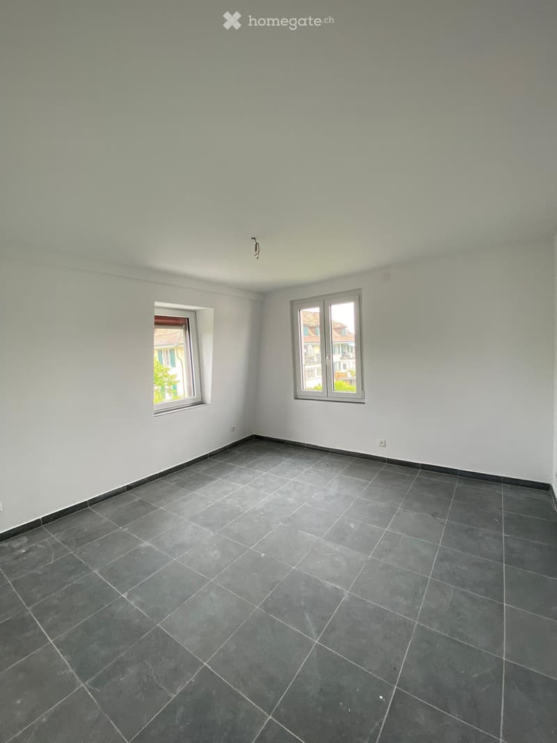 Empty room with grey tiled floor and white walls, featuring two windows with white frames and visible electrical outlets.