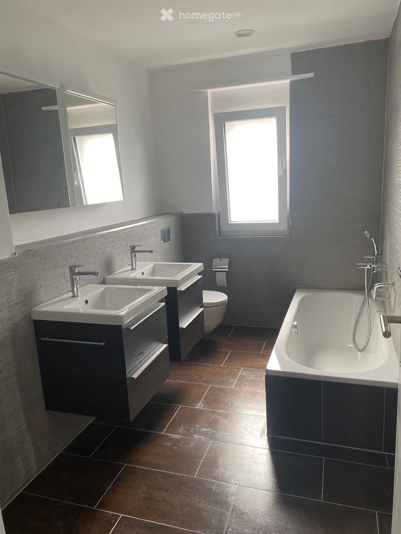Modern bathroom with dark grey tiles, brown tiled floor, double sink vanity, bathtub, and large windows providing natural light.
