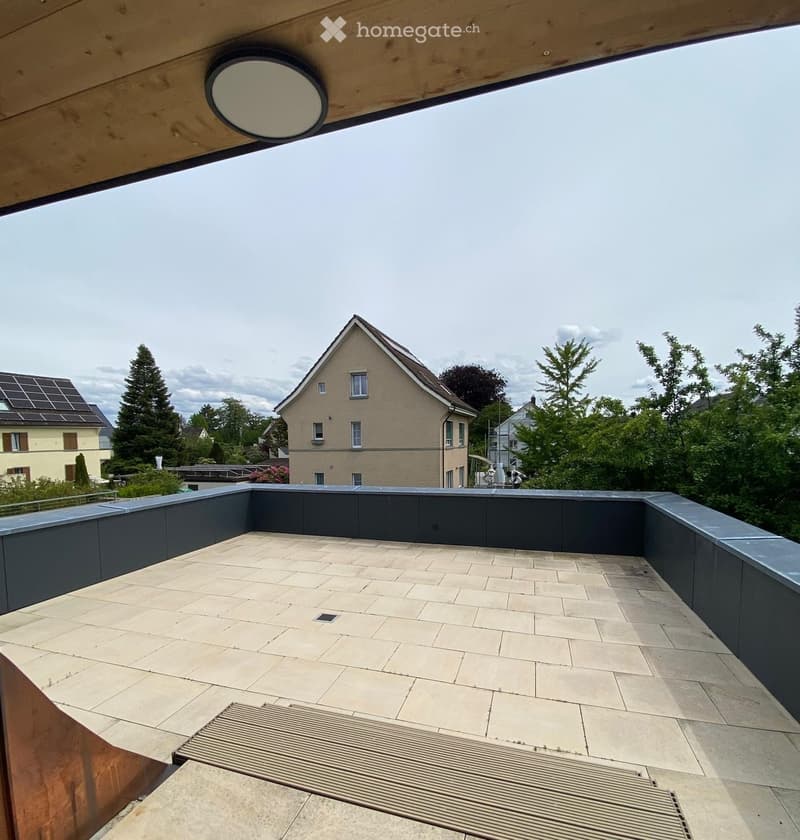 Modern rooftop terrace with beige tiled flooring, dark grey low walls, and a view of surrounding houses and greenery.