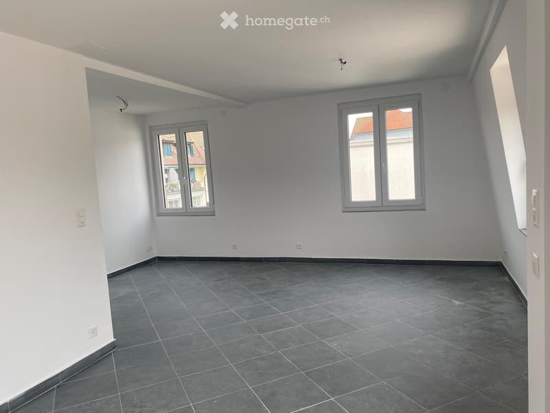 Empty room with grey tiled floor, white walls, and two windows with grey frames. No furniture or equipment present.