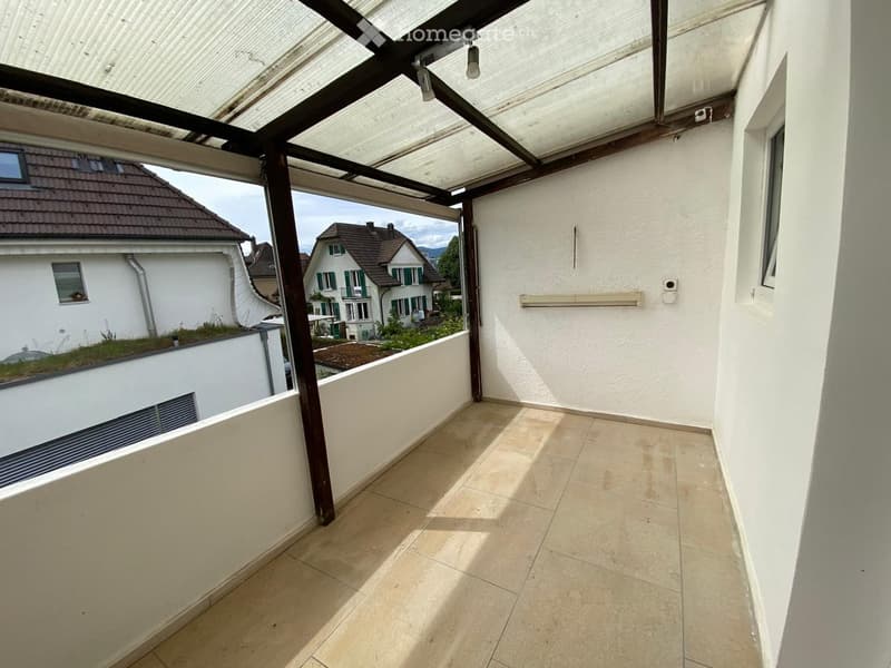 Balcony with tiled floor, white walls, and a covered section with wooden beams and transparent roofing. Offers a view of neighboring houses and greenery.