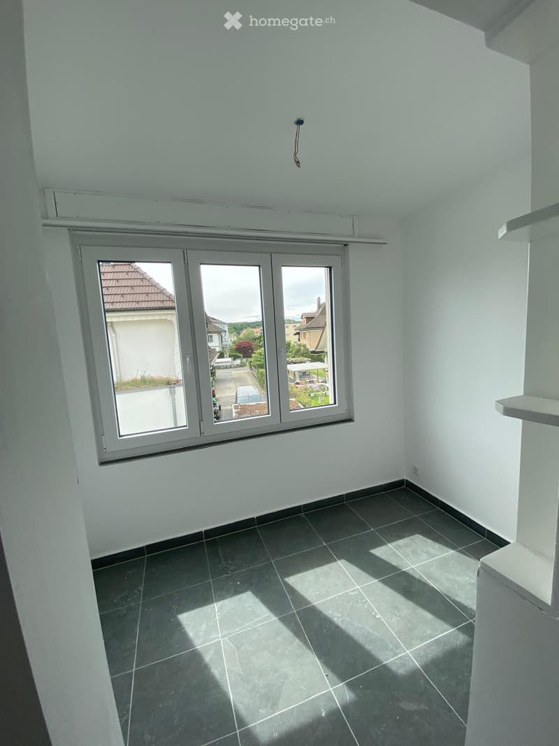 Empty room with white walls, dark tiled floor, and large window offering a view of the outside area.