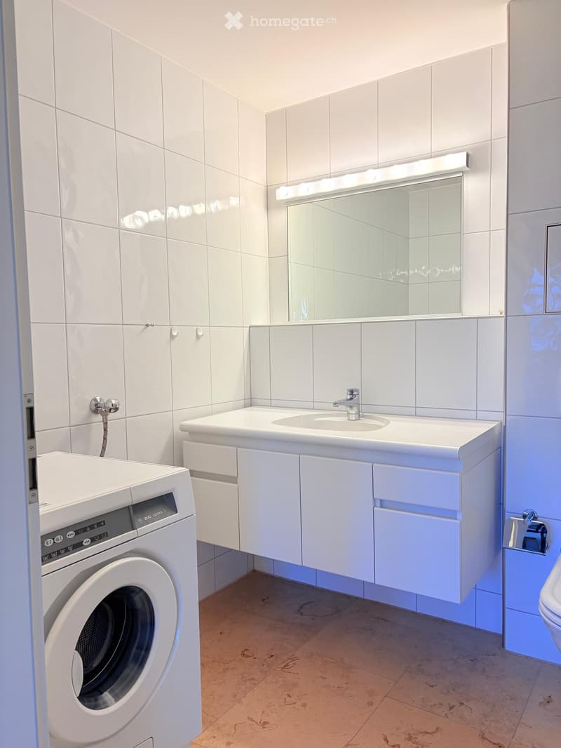 Modern bathroom with white tiles, a sink with a mirror, a washing machine, and a toilet.