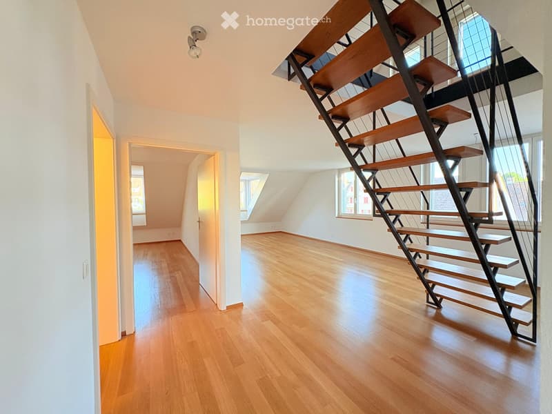 Modern hallway with wooden flooring, white walls, and a contemporary staircase featuring wooden steps and metal railings. Natural light streams in from windows on the right side.