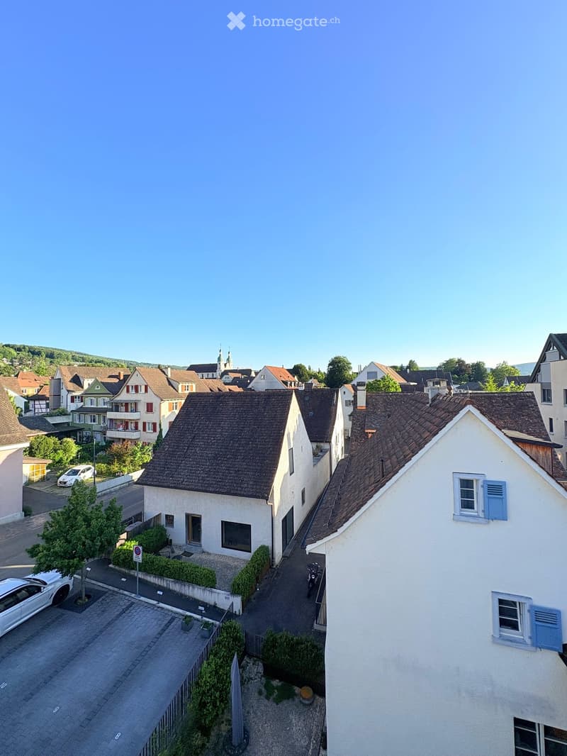 Aerial view of a residential area with multiple houses, gardens, and parked cars. The houses have varied architectural styles and are surrounded by greenery.