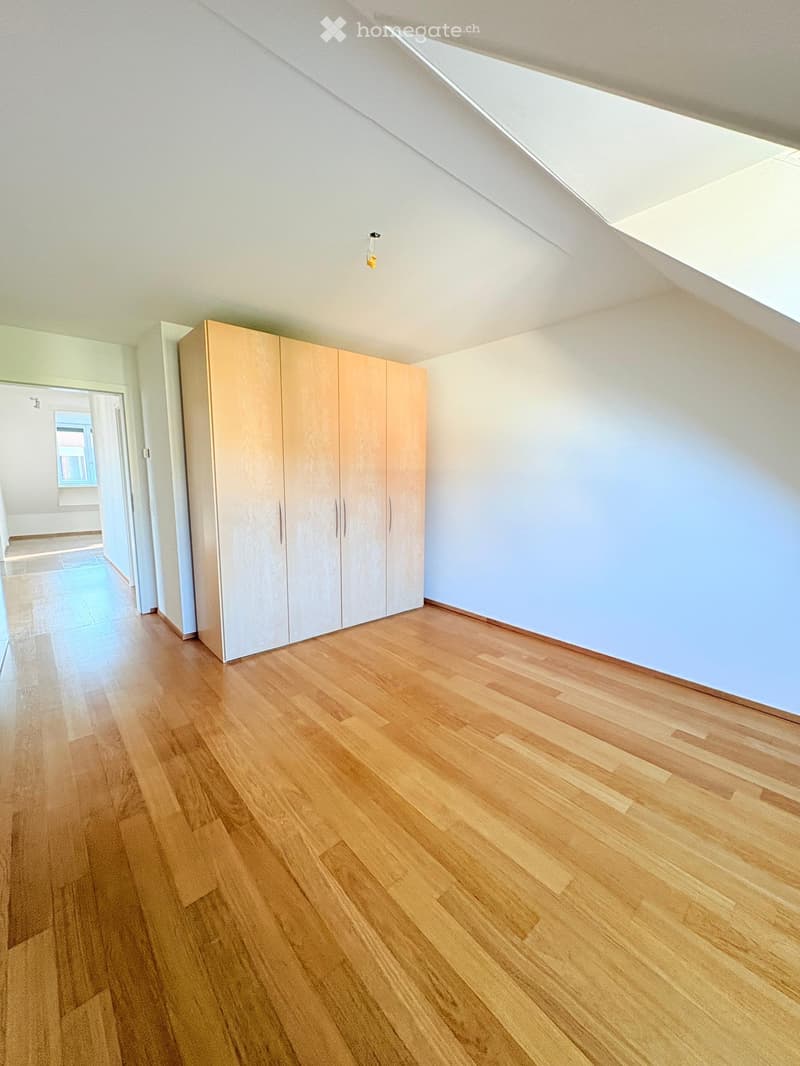 Empty room with wooden flooring, white walls, and built-in closet with sliding doors. Natural light from a skylight and adjacent room.