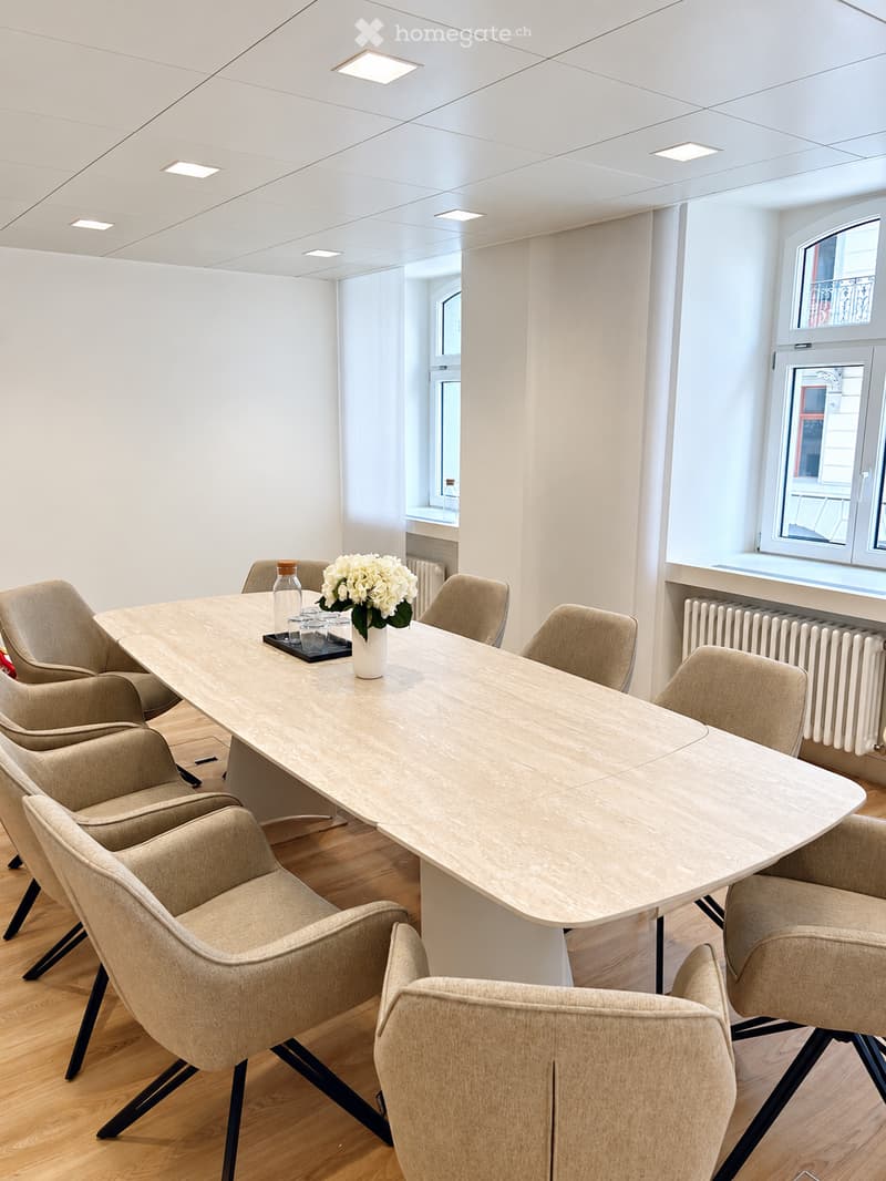 Modern conference room with a long wooden table surrounded by beige chairs, a vase with white flowers, and a bottle on the table.