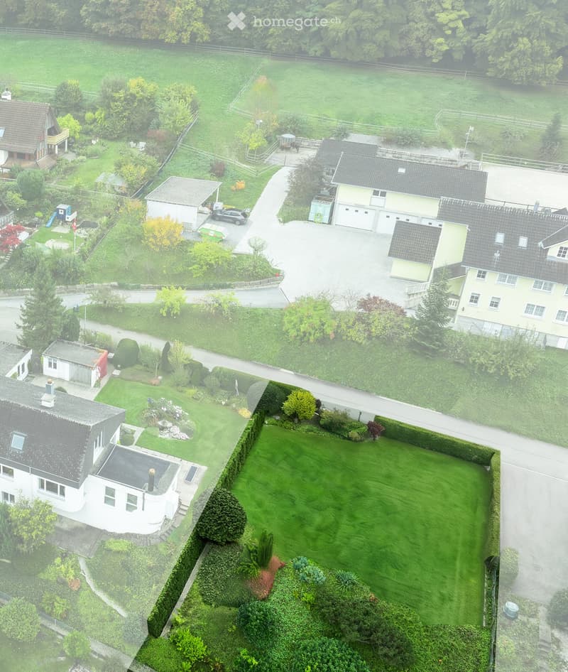 Aerial view of a residential area with multiple houses, well-maintained lawns, gardens, driveways, and parked cars.