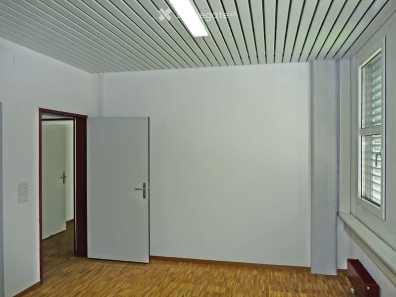 Empty room with white walls, wooden floor, and a single door leading to another space. The room has a window with blinds and a radiator under it.