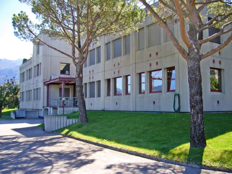 A multi-story building with a modern, minimalist design, featuring large windows and a concrete facade. The building is surrounded by a well-maintained lawn and trees, with a small covered entrance area visible on the left side.