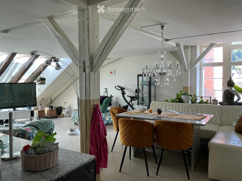Bright, open living space with exposed wooden beams, large windows, and skylights. Features a sofa, dining table with chairs, TV, exercise bike, and decorative items.