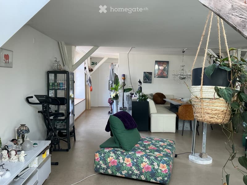 A modern, open-concept living space with a floral-patterned sofa, white ottoman, wicker hanging chair, exercise bike, and decorative items including lamps and picture frames.