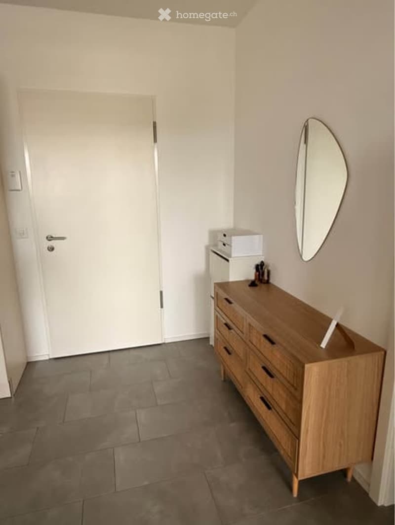 A small, neatly furnished room with a wooden dresser, a round mirror, and a small cabinet. The room has a tiled floor and white walls.