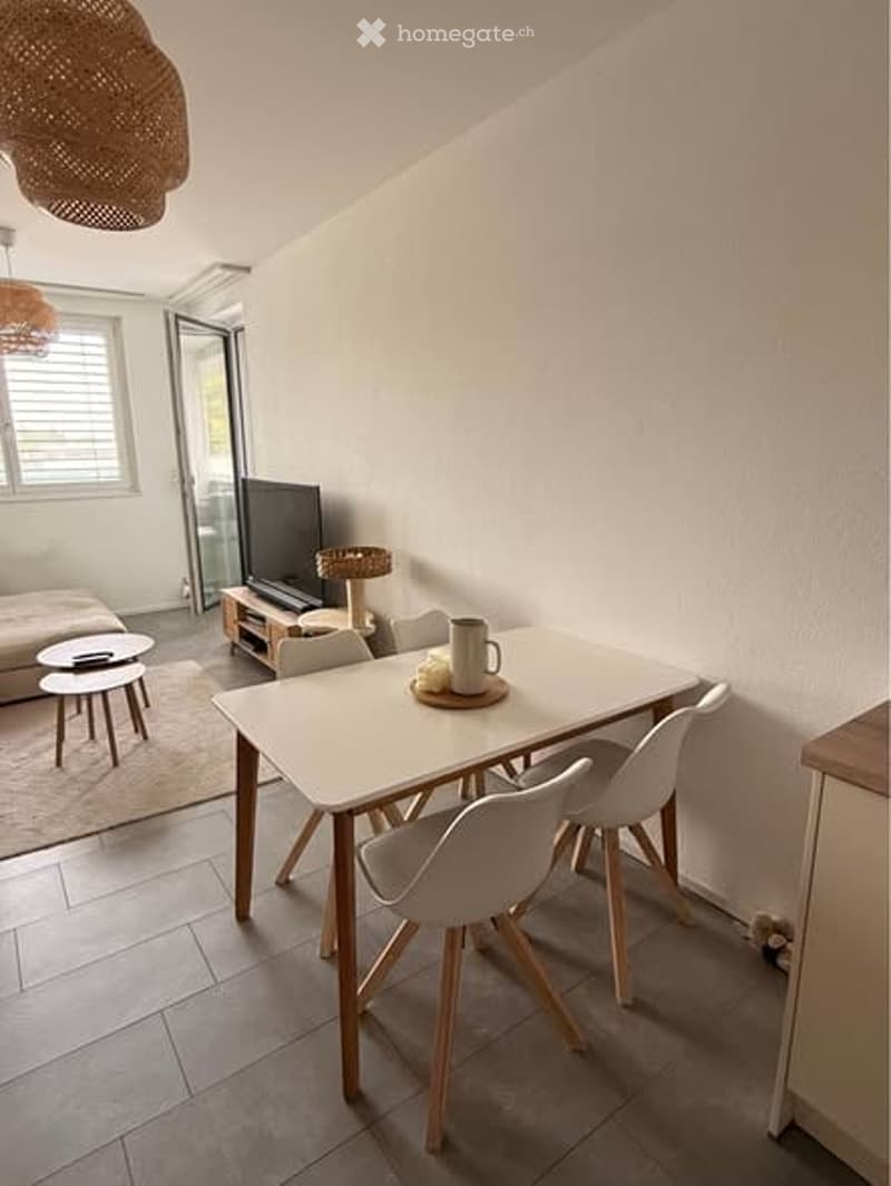 Bright, modern living and dining area with white walls, tiled floor, and large window. Features a sofa, coffee table, dining table with chairs, and a television on a stand.