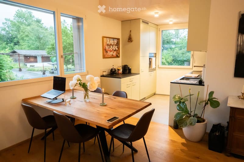 Bright, modern kitchen and dining area with a wooden dining table, chairs, laptop, vase with white flowers, and a candle. The kitchen features white cabinets, a sink, and a coffee maker, with large windows offering a view of greenery outside.