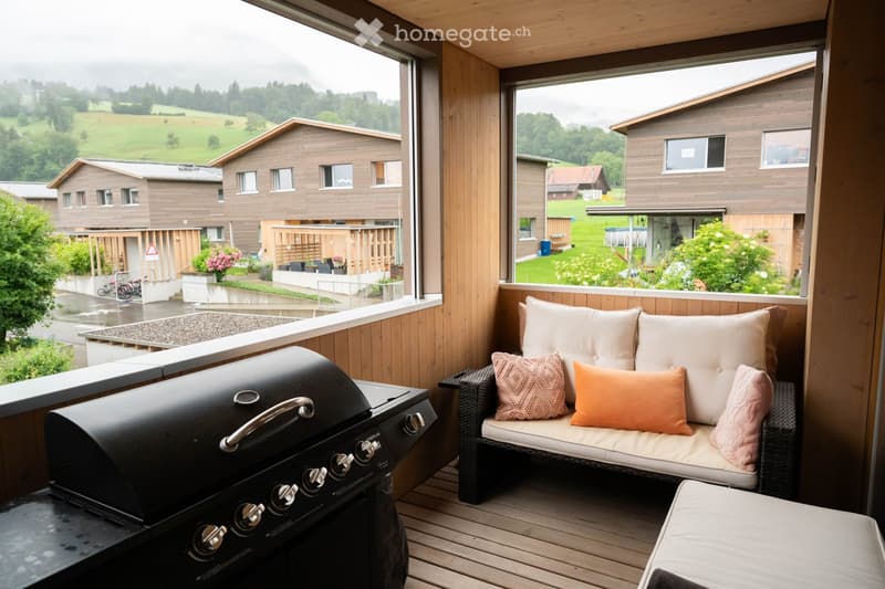A wooden balcony with a wicker sofa, colorful cushions, and a black grill, overlooking a scenic countryside view.