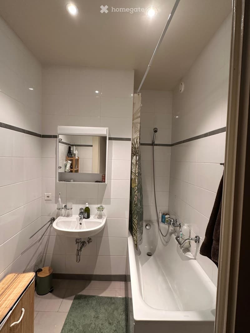 Modern bathroom with white tiles, a sink, bathtub, mirror, and a small wooden cabinet. The space includes a towel, a rug, and a shower curtain with a leafy pattern.