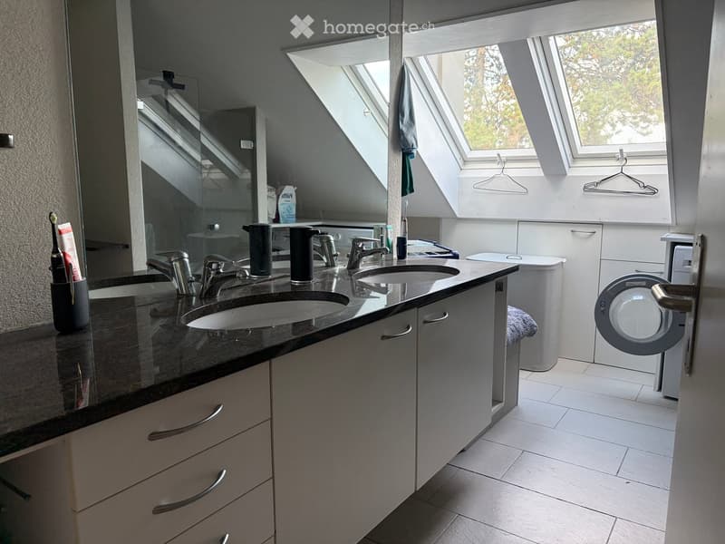 Modern bathroom with double sink, mirrored cabinet, and built-in washing machine. Features skylight windows and white tiled flooring.
