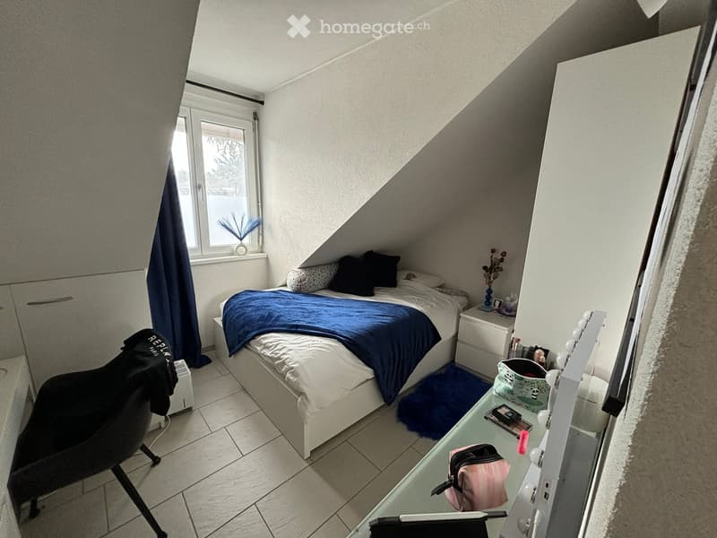 Cozy attic bedroom with a bed, nightstand, mirror, chair, and decorative items. The room features slanted ceilings and a window with a curtain.