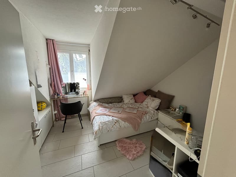 Cozy attic bedroom with slanted ceilings, tiled floor, bed with pillows and blanket, chair, mirror, small table, cabinet, and a round pink rug.
