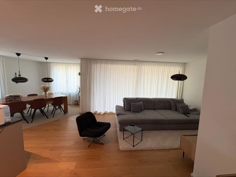 Modern living and dining area with a grey sectional sofa, black coffee table, black armchair, wooden dining table with four chairs, pendant lights, and a vase with dried flowers on the dining table.