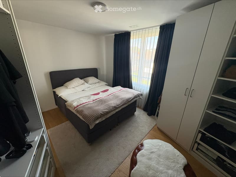 A modern bedroom with a double bed, white bedding, a large wardrobe, and a window with dark curtains.