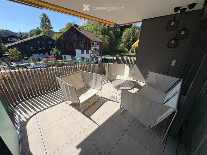 Balcony with modern white chairs and a small round table, overlooking a scenic area with houses and trees.