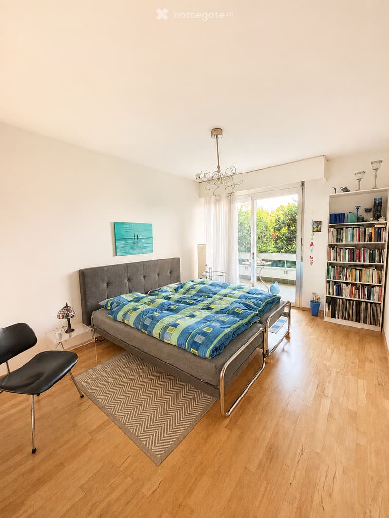 Bright bedroom with wooden flooring, a grey upholstered bed with blue and green bedding, a black chair, a small table with a lamp, a bookshelf filled with books and decorative items, and a painting on the wall. A large window with sheer curtains leads to