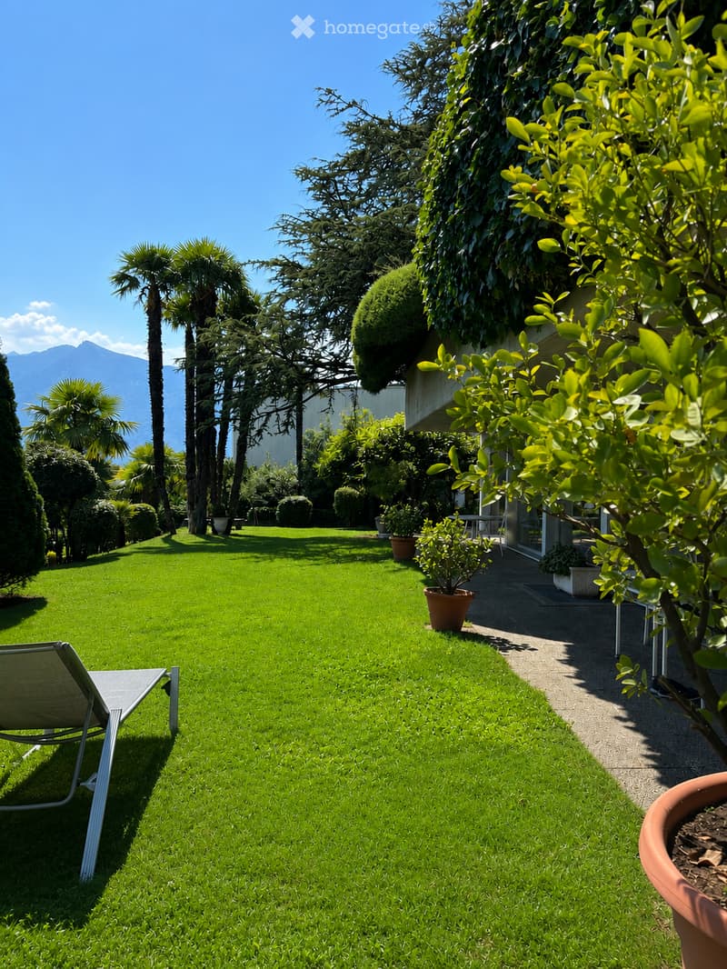 Well-maintained garden with green lawn, lounge chair, potted plants, and trees, with a view of mountains in the background.
